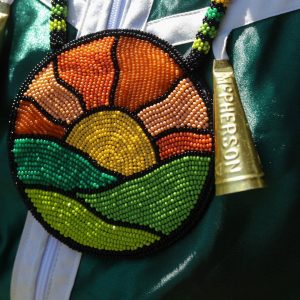 Jingle dress dancer Lauren Porter made her medallion by hand using glass beads. The jingle dress style originated after contact with settlers, and incorporates materials that were adopted by First Nations to reflect and express their worldview. Hanging beside the medallion is a jingle made from the lid of a Copenhagen snuff tobacco tin.