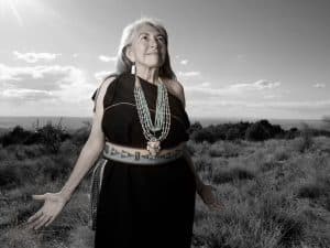 A Native woman photographed by Matika Wilbur. She looks up at the sky with her hands extended at her sides.Dr. Mary Evelyn Belgarde, Pueblo of Isleta and Ohkay Owingeh.Matika Wilbur