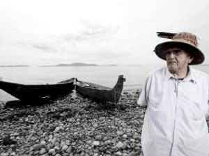 Chief Bill James, Lummi Nation. A man poses for a portrait in front of a canoe and water.Chief Bill James, Lummi Nation.Matika Wilbur