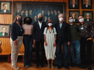 Seven people wearing masks stand in front of a wall of portraits of white men