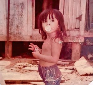 A toddler with a pacifier in her mouth stands outside a rickety wooden building.