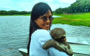 A smiling woman stands by a river holding a sloth.
