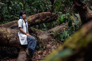 A woman with Indigenous face markings and wearing a western-style white coat and stethoscope sits on a fallen log in a forest.