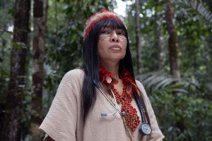 A woman wearing beads and a stethoscope around her neck stands in the forest. She has a feather headband and a facial tattoo
